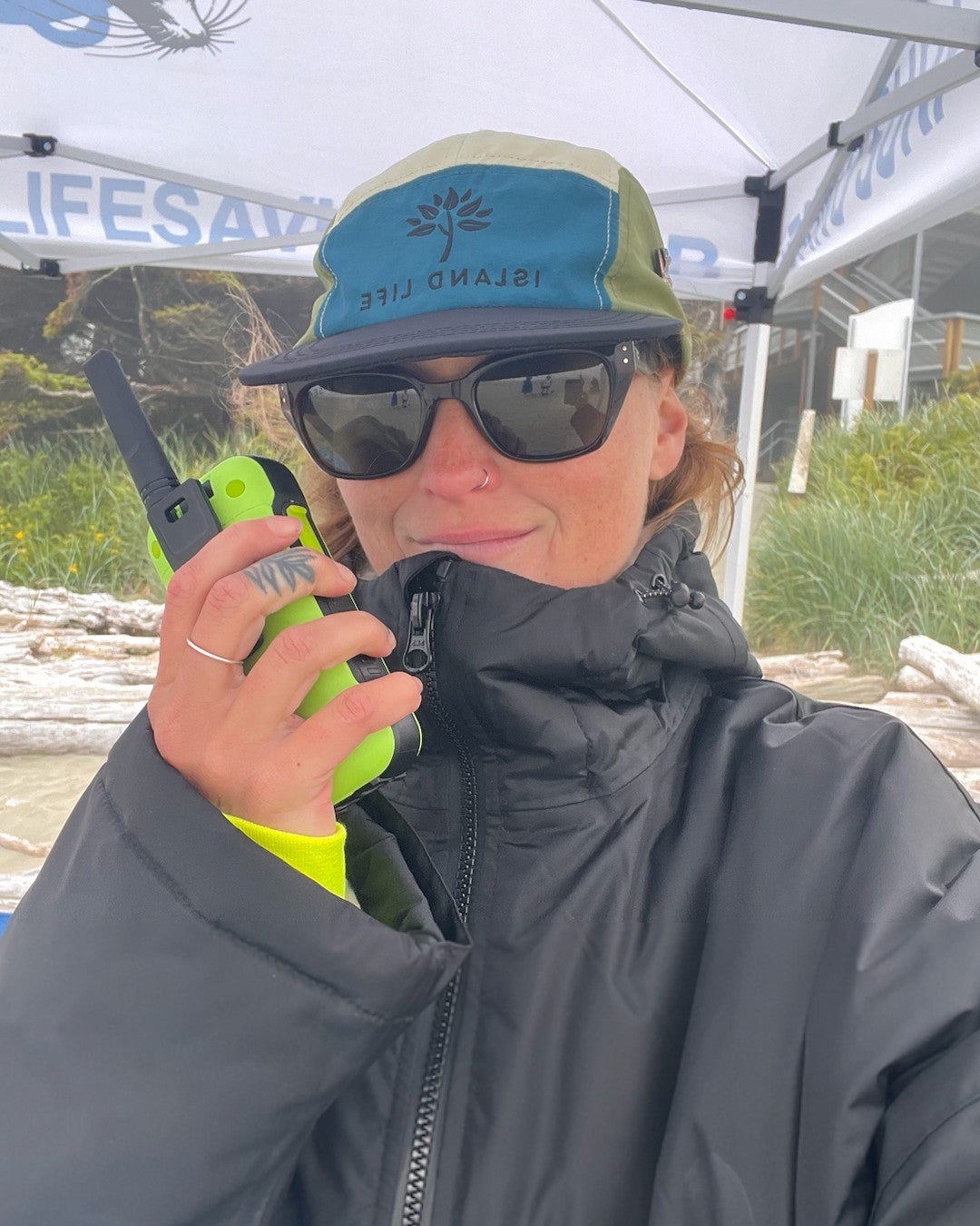 Person wearing a cap and sunglasses, holding a walkie-talkie with a 'Lifeguard' background.