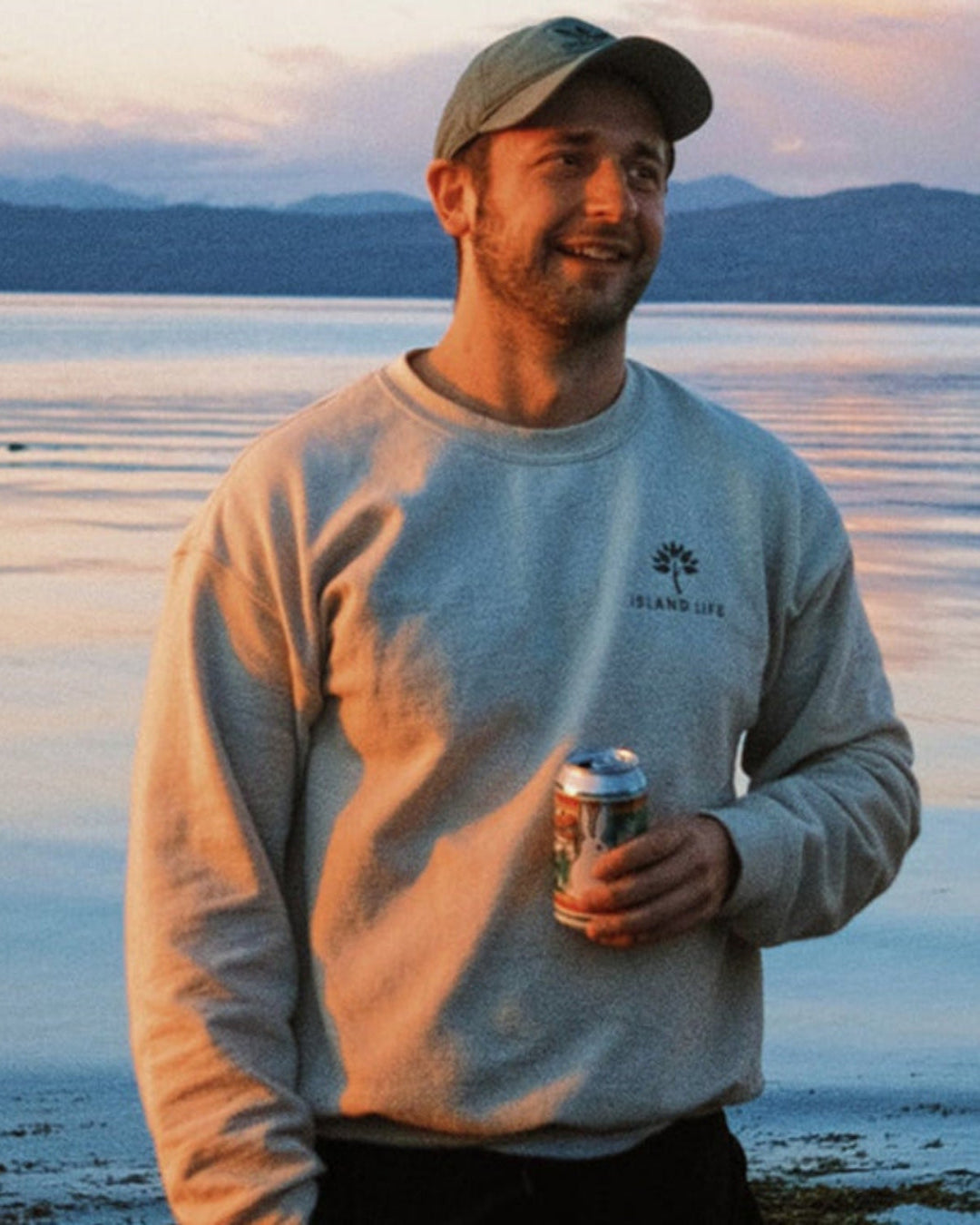 Man holding a can by a body of water with mountains in the background